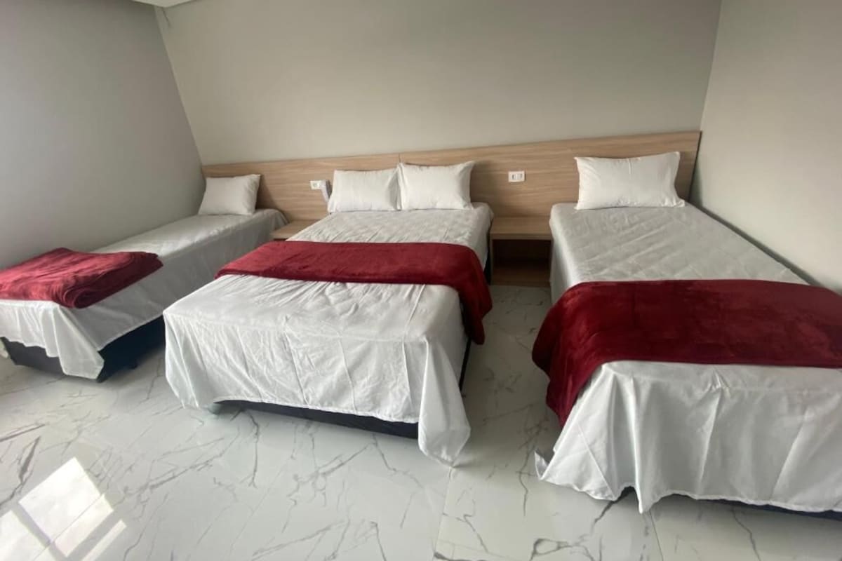 Habitación de hotel con tres camas dispuestas en fila, cabecera de madera y suelo de mármol.