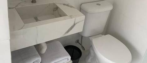 Un baño con lavamanos de mármol, inodoro y estante de vidrio sobre el lavamanos.