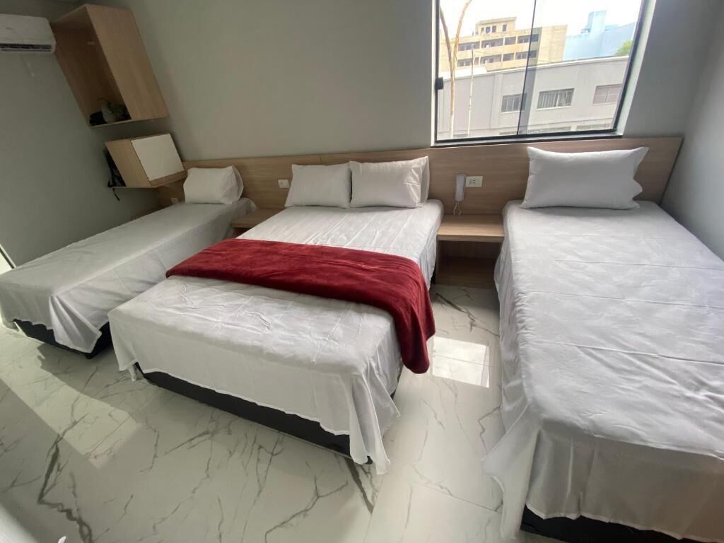Habitación de hotel con tres camas, una manta roja sobre la cama central, una ventana con vistas a edificios y un suelo de mármol.