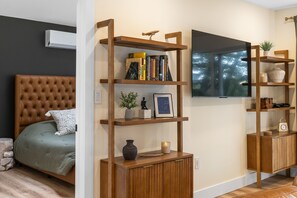 Une chambre à coucher avec une tête de lit capitonnée, un lit agrémenté d’un coussin à motifs, une bibliothèque en bois contenant des livres et des objets décoratifs, un téléviseur fixé au mur et une table de chevet ornée d’un vase et d’un tableau encadré.