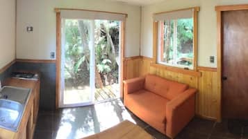 A compact room with a kitchenette, a sofa, and a sliding glass door leading to a natural outdoor area.