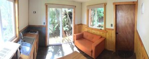 A compact room with a kitchenette, a sofa, and a sliding glass door leading to a natural outdoor area.