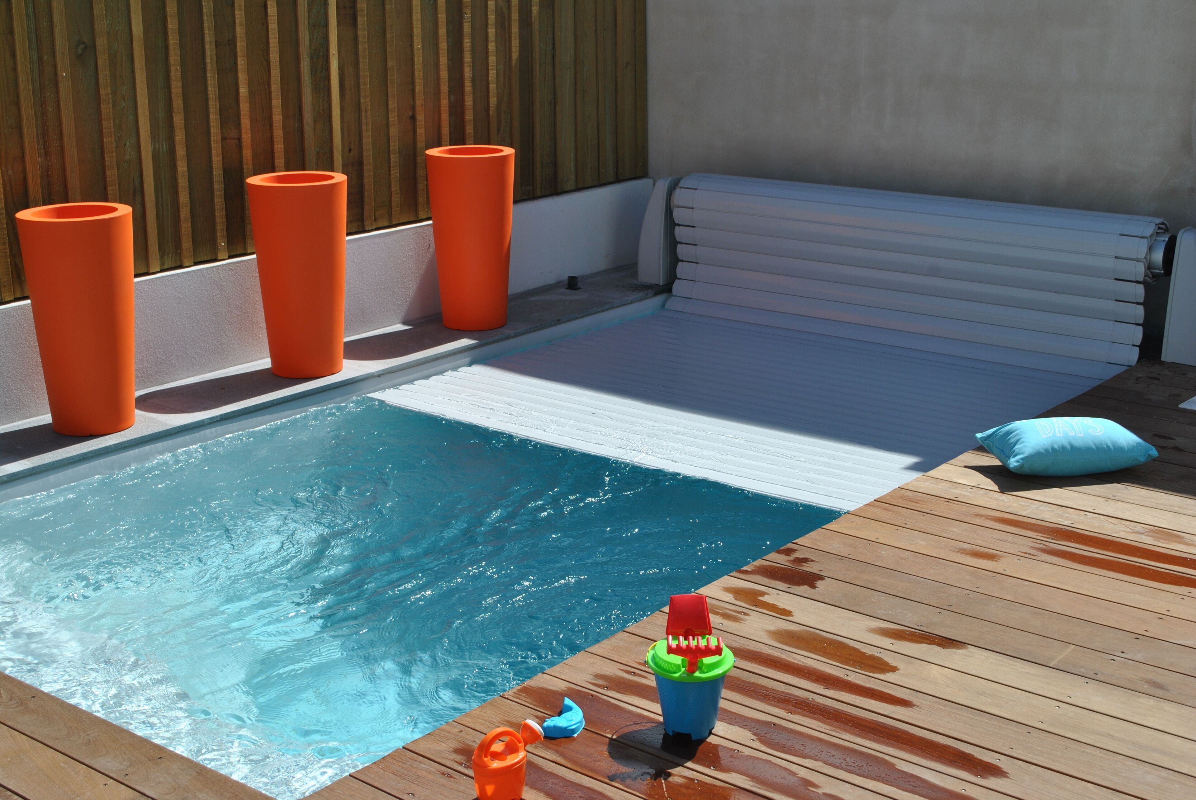 Una piscina con un solarium in legno, una zona relax con un divano bianco e oggetti cilindrici arancioni.