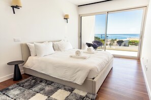 Ein Schlafzimmer mit einem großen Bett, einem kleinen Tisch, einem Teppich und Meerblick durch eine Schiebetür.