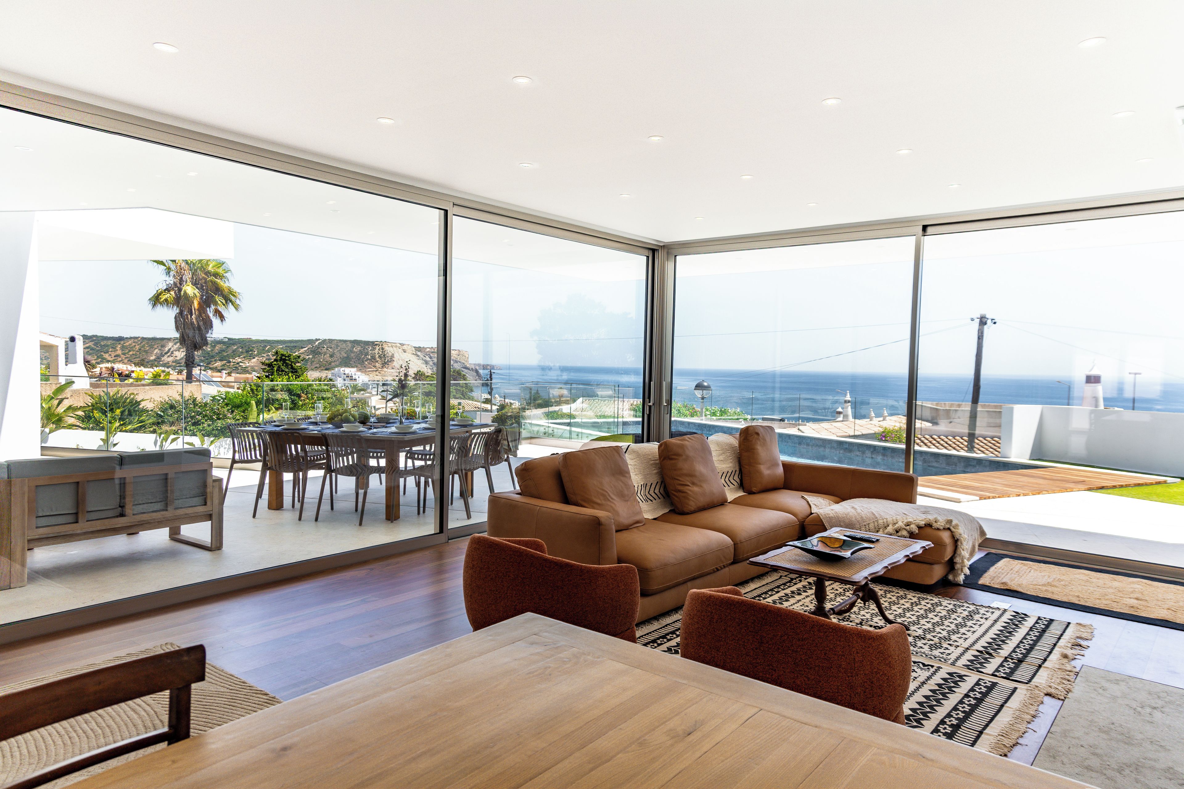 A modern living room with a large glass wall, a wooden dining table, and a brown sofa.