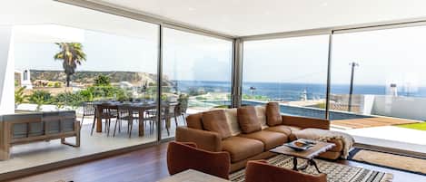 A modern living room with a large glass wall, a wooden dining table, and a brown sofa.