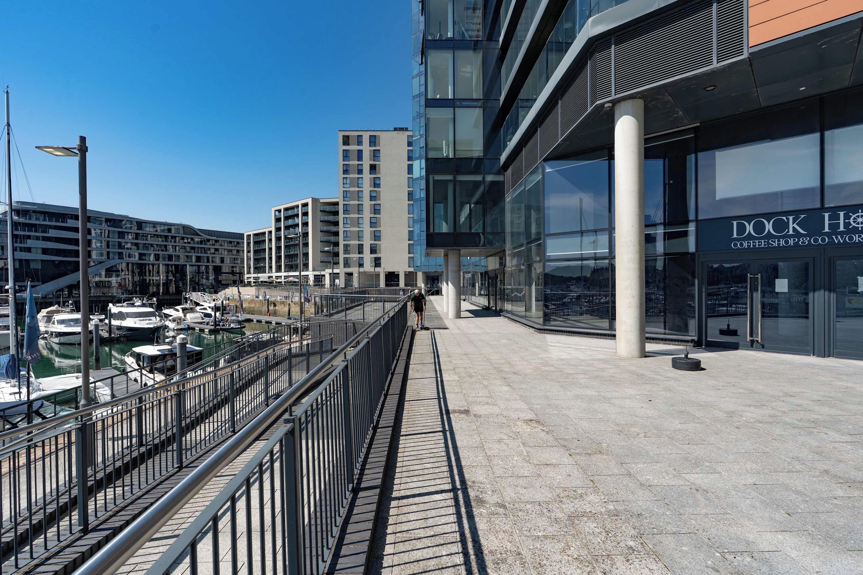 Een waterkantpromenade met een koffiezaak genaamd 'DOCK H' en een modern gebouw met balkons.