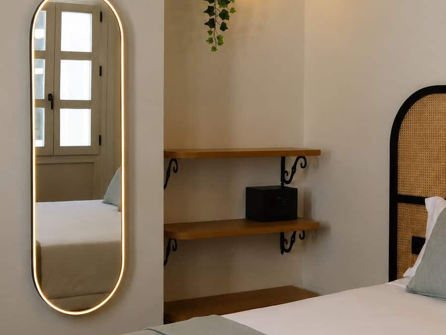 Quarto com cama, mesa de cabeceira, espelho, prateleira na parede com uma planta e luminária de parede.