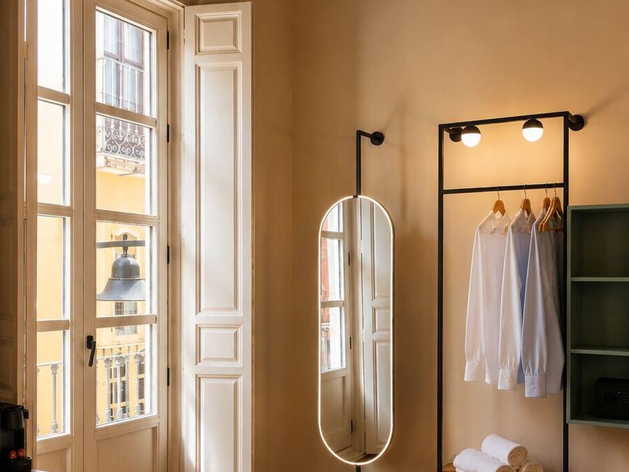 Un dormitorio con techo de madera, un espejo grande, un armario con ropa colgada y una cama con colcha color beige.