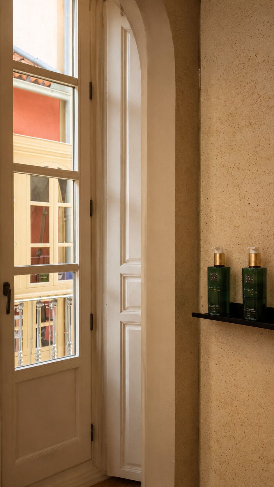 Uma porta branca, uma janela com vista para um prédio e uma prateleira com duas garrafas verdes.