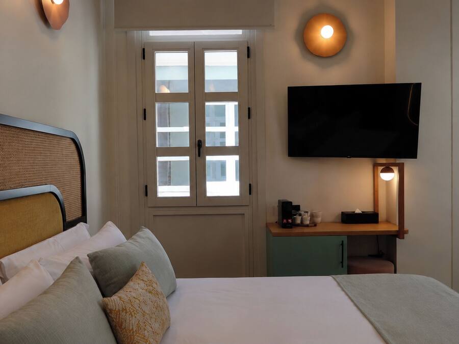 Una habitación de hotel con cama, televisión montada en la pared, ventana con persianas y mesita de noche con lámpara.