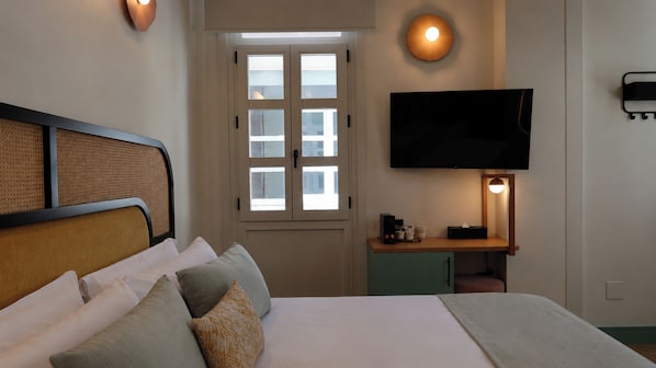 Una habitación de hotel con cama, televisión montada en la pared, ventana con persianas y mesita de noche con lámpara.