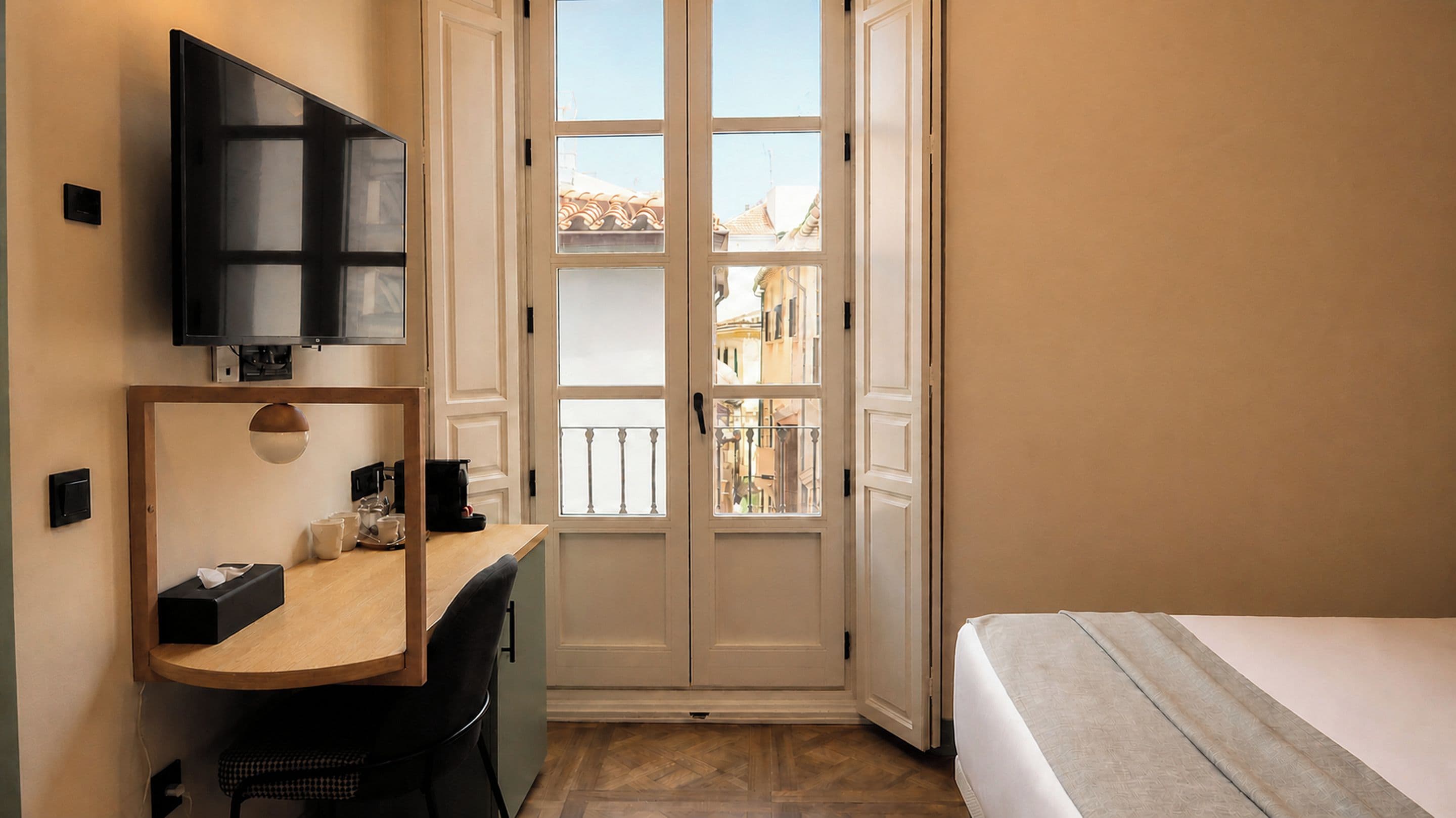 Habitación de hotel con cama, escritorio, silla, televisor y ventana con vista a edificios.