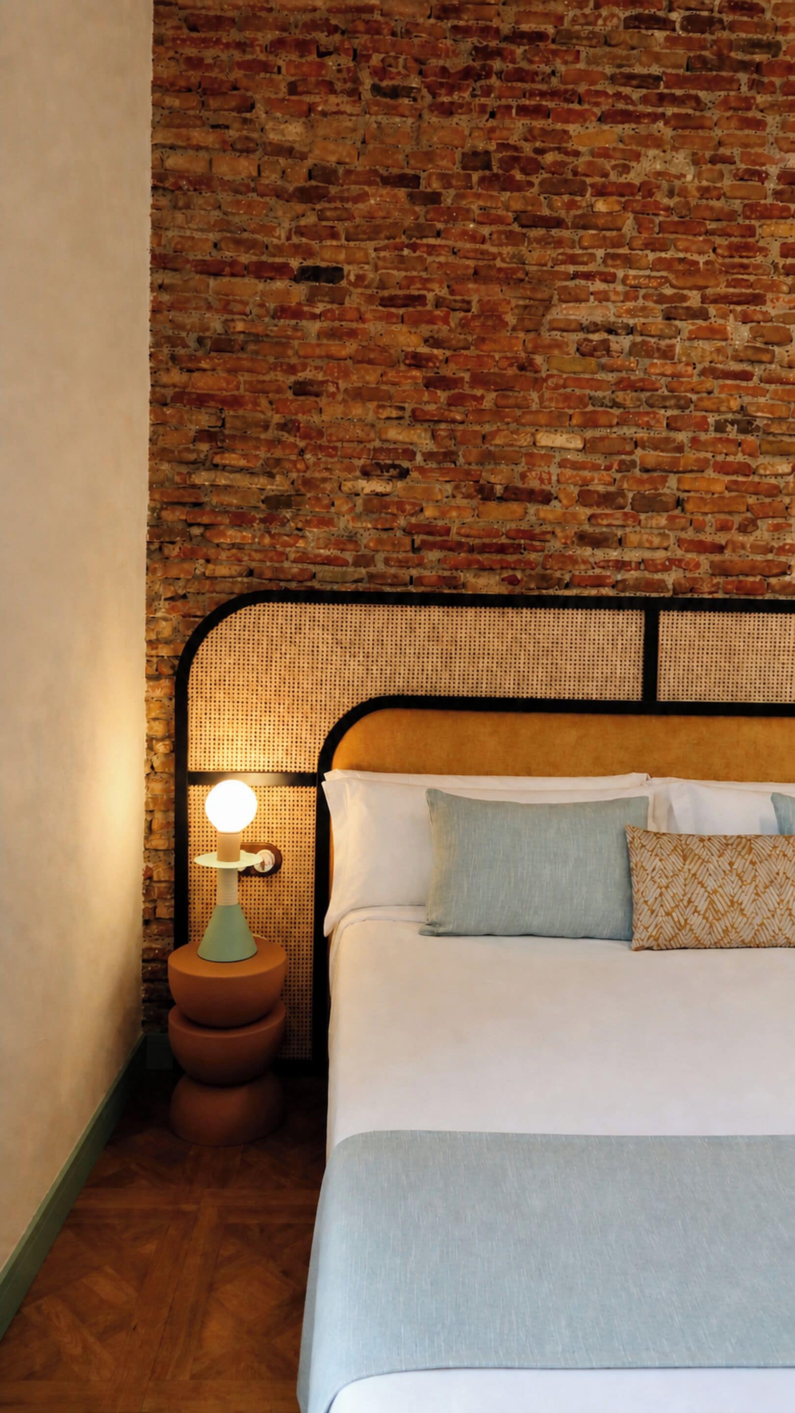 Una cama con cabecera apoyada en una pared de ladrillo, una mesita de noche con lámpara y un cojín estampado.