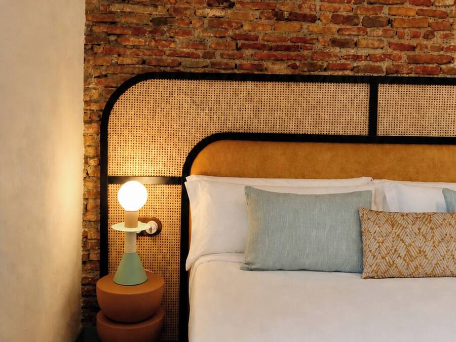 Una cama con cabecera apoyada en una pared de ladrillo, una mesita de noche con lámpara y un cojín estampado.