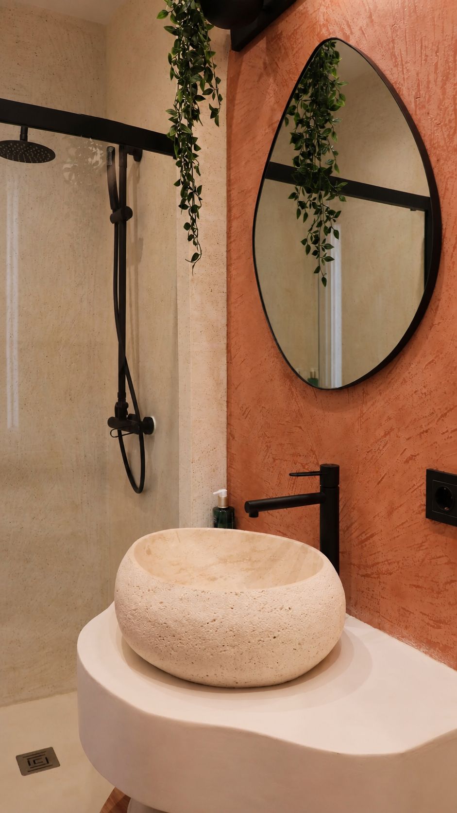 A bathroom with a round stone sink, a black faucet, a handheld showerhead, and a mirror with a plant hanging above it.