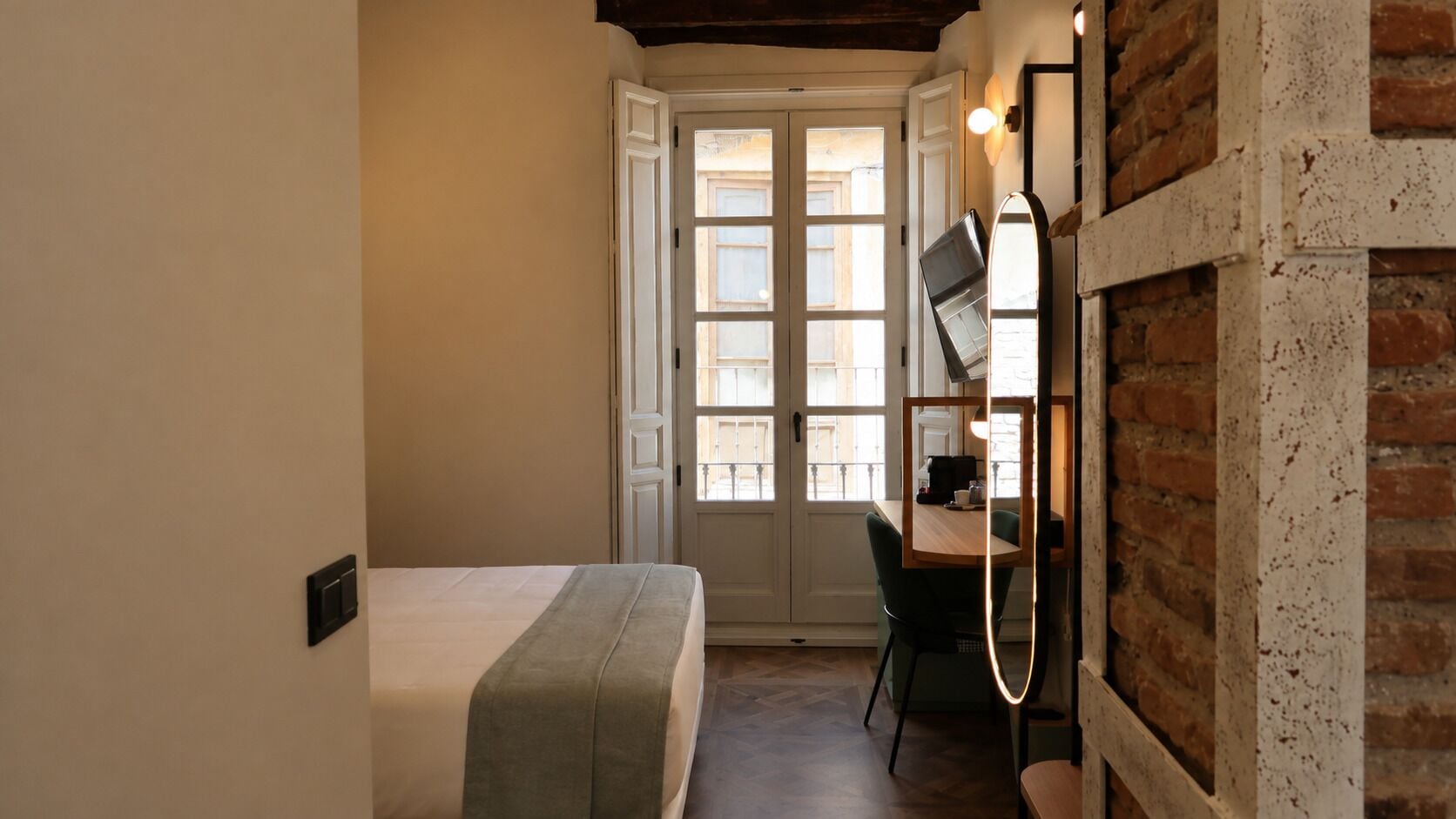 Een slaapkamer met een bed, een bureau en een stoel, een deur naar een buitenbalkon, en een bakstenen muur.
