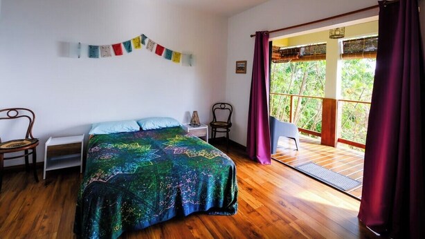 Quarto com cama, cadeira, mesa de cabeceira, janela com cortinas e vista para o verde lá fora.