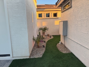 A narrow pathway with artificial grass, surrounded by white textured walls and a small palm tree. - Spacious 5-bedroom house in marvelous Maricopa with AC (Maricopa)
