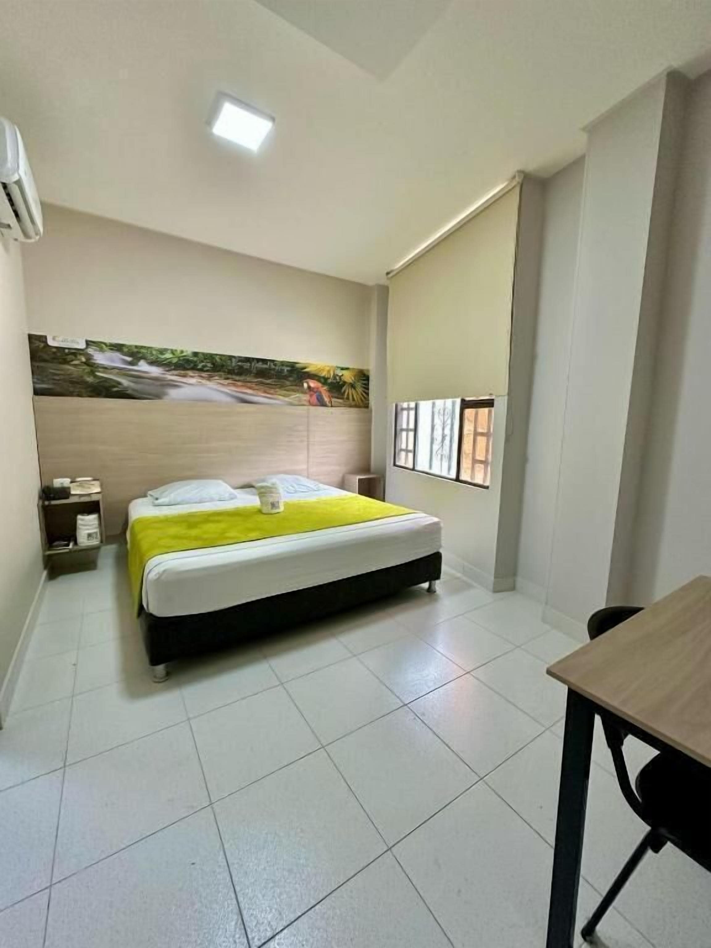 A hotel room with a bed, a desk, a chair, a nightstand, a wall-mounted air conditioner, and a window with blinds.