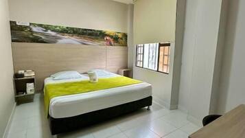 A hotel room with a bed, a desk, a chair, a nightstand, a wall-mounted air conditioner, and a window with blinds.