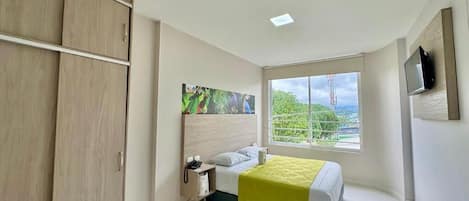 Quarto de hotel com cama, escrivaninha, armário, janela com vista e televisão presa à parede.
