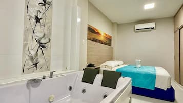 A hotel room with a large bathtub, a bed with a blue bedspread, a painting on the wall, and an air conditioning unit.