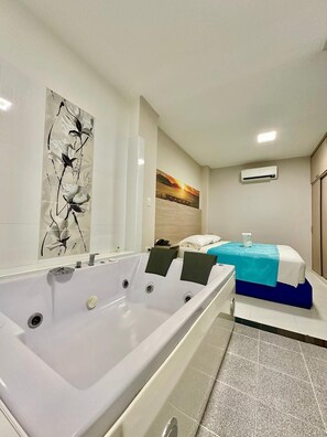 Quarto de hotel com uma banheira grande, cama com colcha azul, um quadro na parede e um aparelho de ar condicionado.