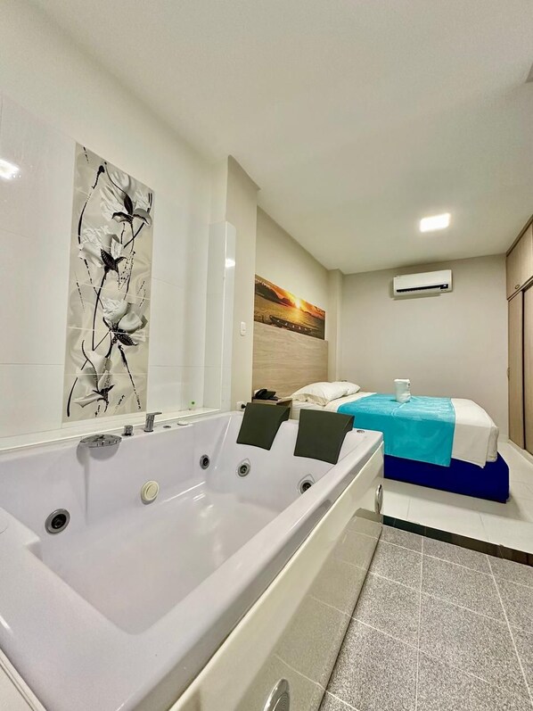 Habitación de hotel con una bañera grande, cama con cubrecama azul, un cuadro en la pared y un equipo de aire acondicionado.