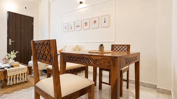 A dining area with a wooden table and chairs, a white wall with framed artwork, and a door in the background.