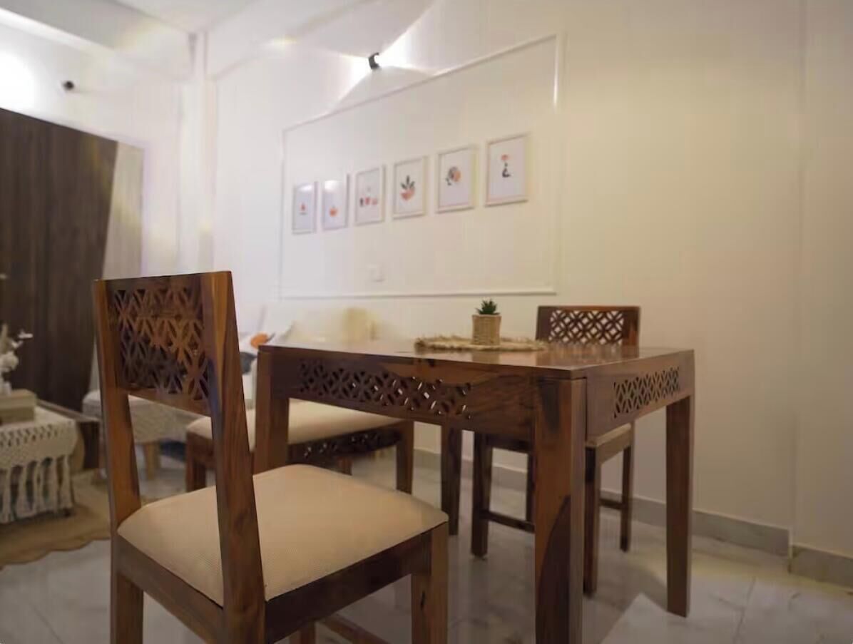 A dining area with a wooden table and chairs, a white wall with framed pictures, and a small potted plant on the table.