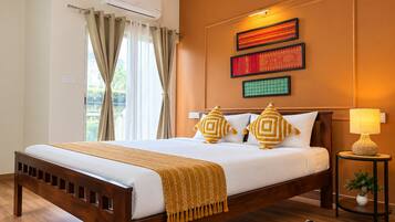 Habitación de hotel con una cama de madera, cortinas color beige, aire acondicionado montado en la pared y una mesita con una lámpara y una planta.