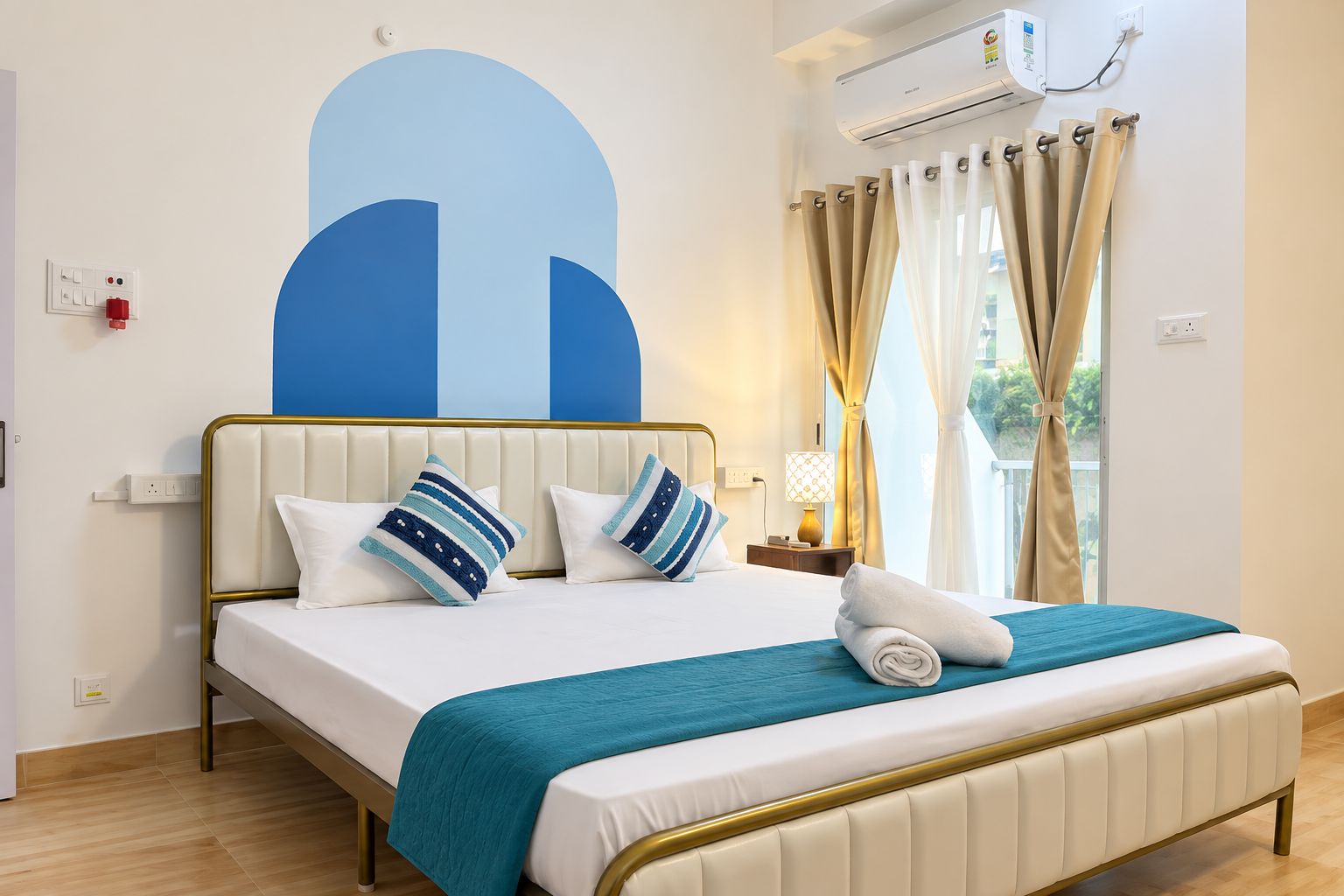 A hotel room with a bed, a headboard, a bedside table with a lamp, a window with curtains, and an air conditioning unit.