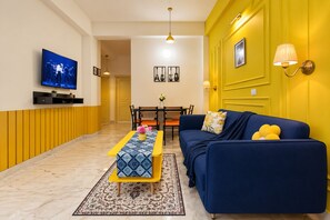 A modern living room with a blue sofa, yellow walls, a TV mounted on the wall, and a dining area with a table and chairs.