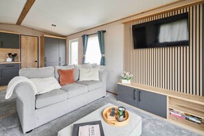 A modern living room with a grey sofa, wooden cabinets, a flat-screen TV, and a tray with books and a remote. - Host & Stay | Wilderness Wood Lodge (Pickering)