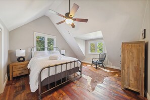 Ein Schlafzimmer mit schrägem Dach, einem Bett mit Metallkopfteil, einer Holzkommode, einem Stuhl und einem Kuhfellteppich.