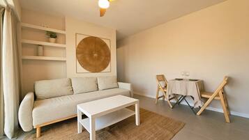 A living room with a sofa, a coffee table, a dining table with chairs, a ceiling fan, and a large wall art piece.