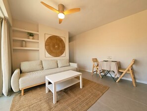 A living room with a sofa, a coffee table, a dining table with chairs, a ceiling fan, and a large wall art piece. - ‘Ocean View’ Apartment with Sea Views, Balcony, and Wi-Fi (Sant Feliu de Guíxols)