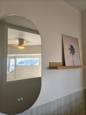 A bathroom with a large mirror, a ceiling fan, a framed picture on the wall, and a window with a view of the sea.