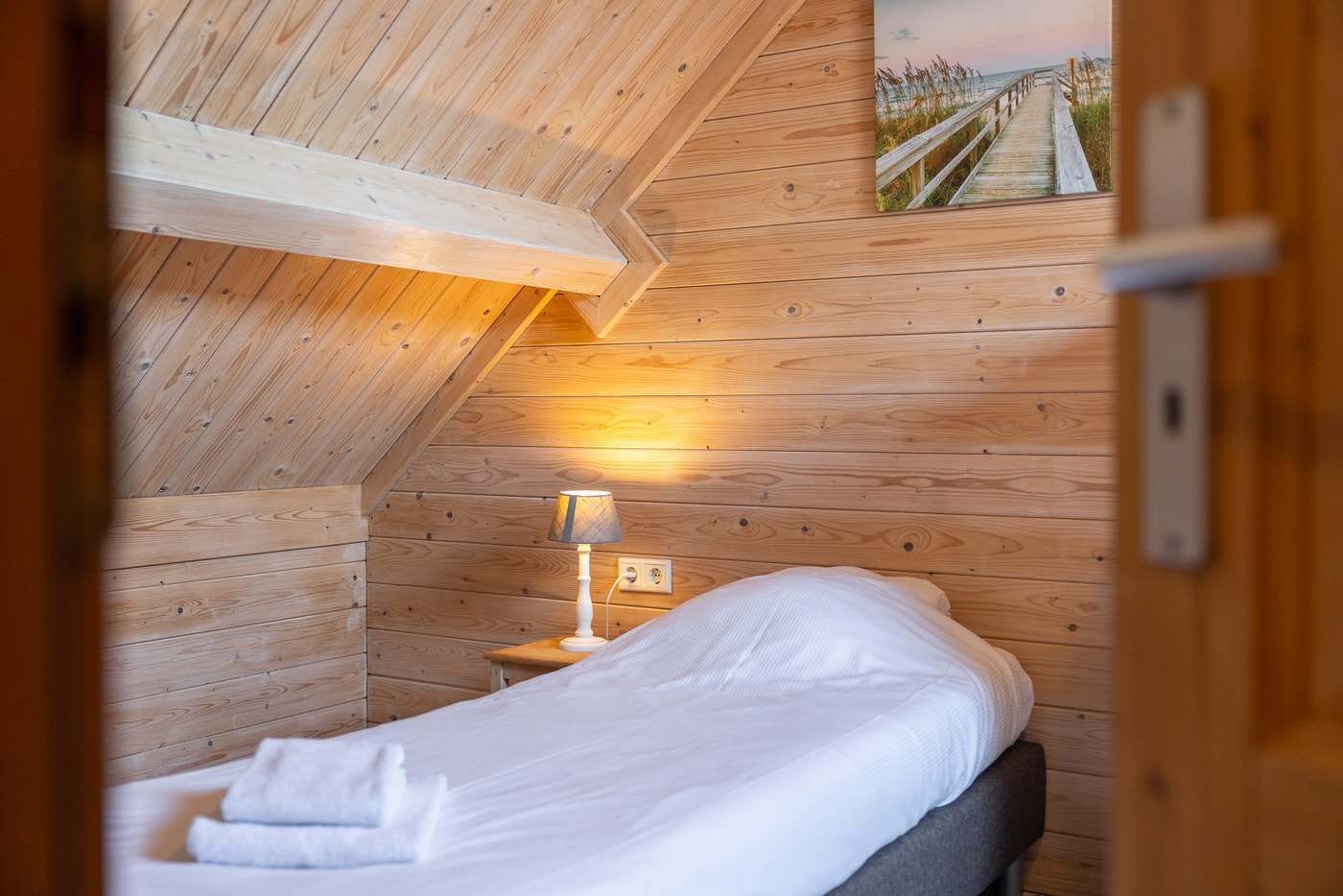 Une pièce aux murs lambrissés, comprenant un lit, une table de chevet, une lampe et un tableau accroché au mur.