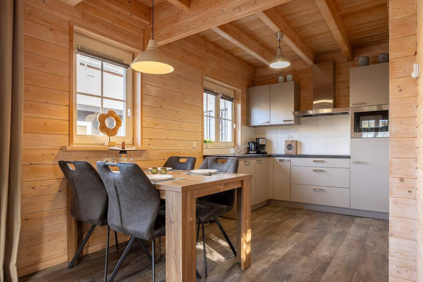 A wooden interior with a dining table, chairs, a kitchen area, and a window with a decorative wreath.
