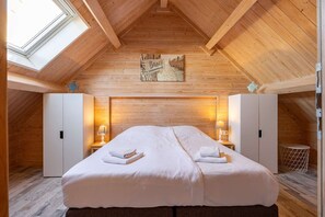 Ein Schlafzimmer mit schrägverlaufender Decke, Holzwänden, einem Bett mit weißen Bettwäsche, zwei Nachttischlampen und einem Gemälde über dem Bett.