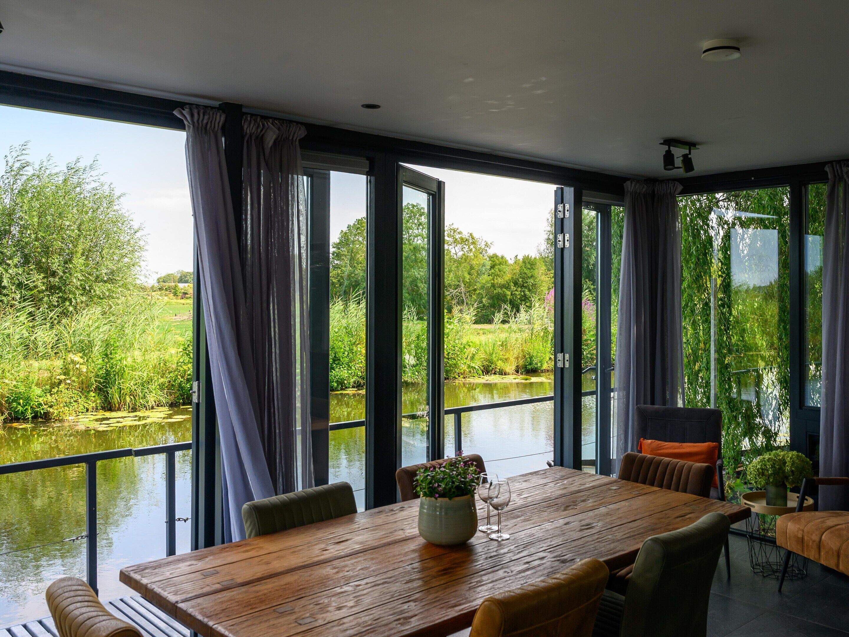 Un espace repas avec une table et des chaises en bois, de grandes fenêtres avec des rideaux, et une vue sur une étendue d’eau et la verdure.