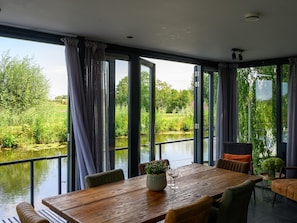 A dining area with a wooden table and chairs, large windows with curtains, and a view of a body of water and greenery. - Waterfront Villa With Picturesque Views (Hekendorp)