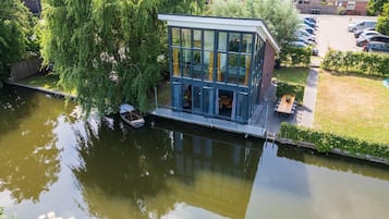 Ein modernes Hausboot mit Glasfassade, an einem Kanal angedockt, umgeben von Grünflächen und Wohngebäuden.