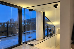 Ein modernes Interieur mit großen Glasüren, die zu einer verschneiten Außenanlage führen, einer Treppe mit Geländer und einem minimalistischen Design.