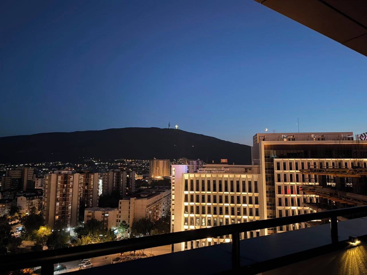 A cityscape at dusk with illuminated buildings and a mountain in the background.