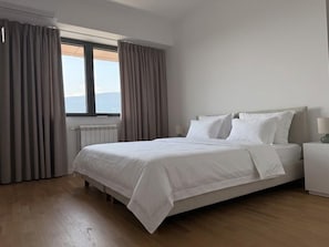 Un dormitorio con cama, mesita de noche, ventana con cortinas y vista al mar.