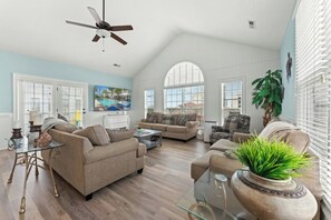 Ein geräumiges Wohnzimmer mit hohen Decken, großen Fenstern und einem Deckenventilator.
