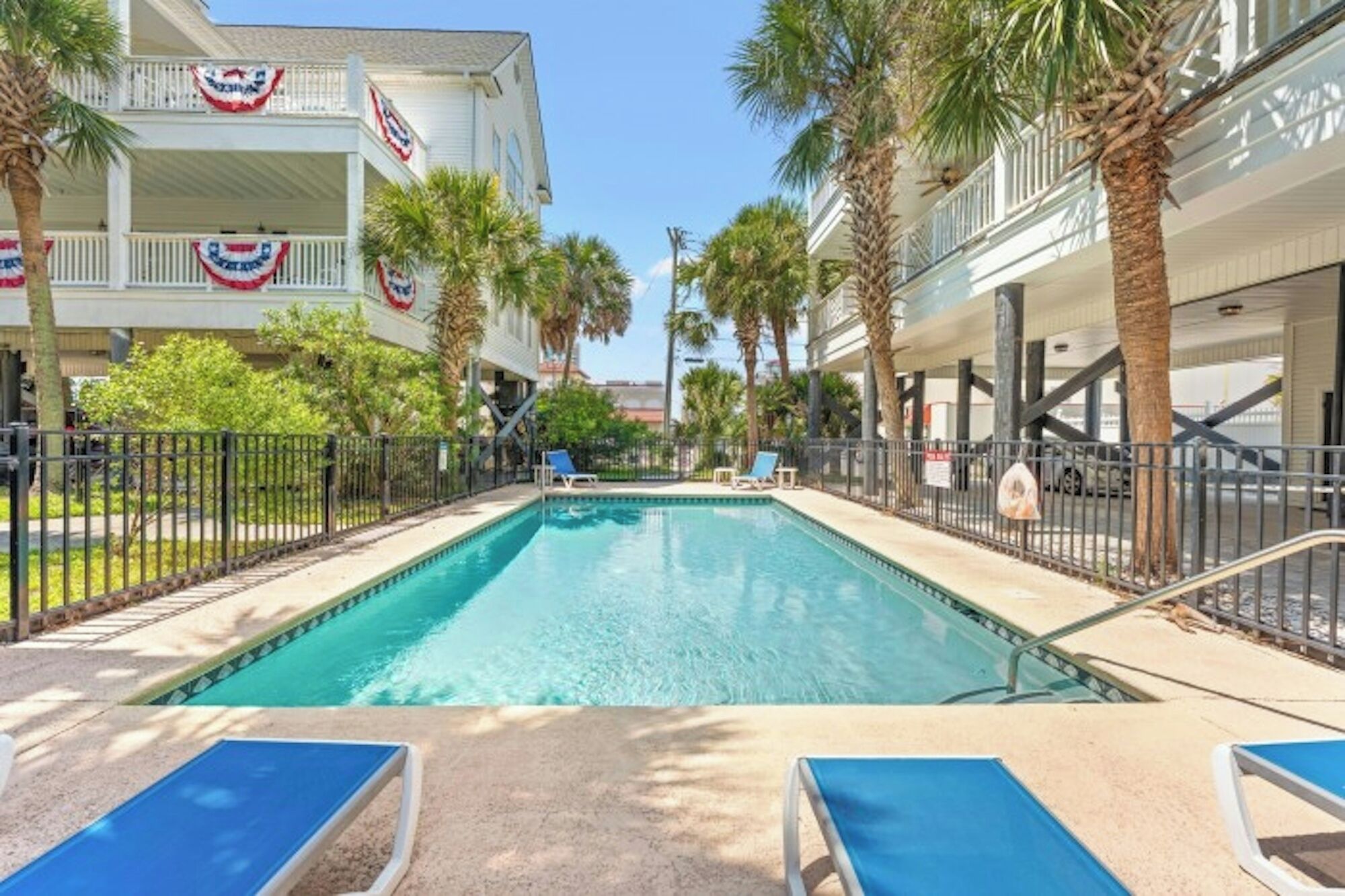 A swimming pool with lounge chairs, palm trees, and a building with American flags.